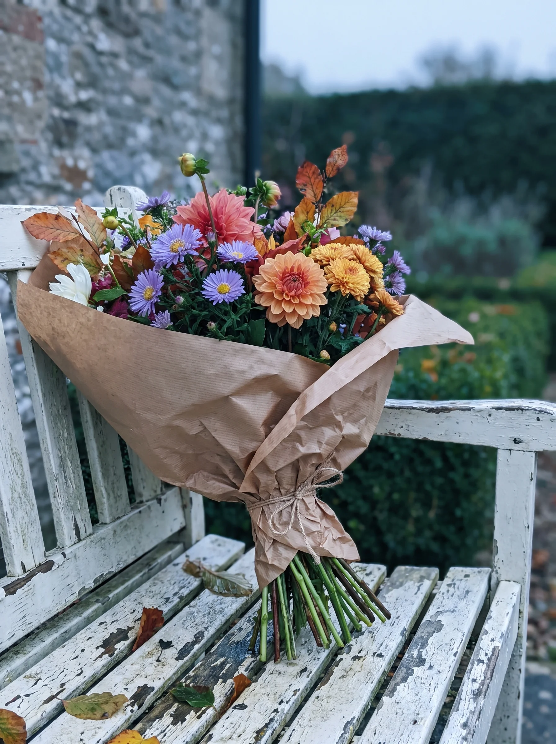 A mixed bunch of dahlias, rudbeckia, scabious and rosehips wrapped in brown paper and tied with string on a white bench